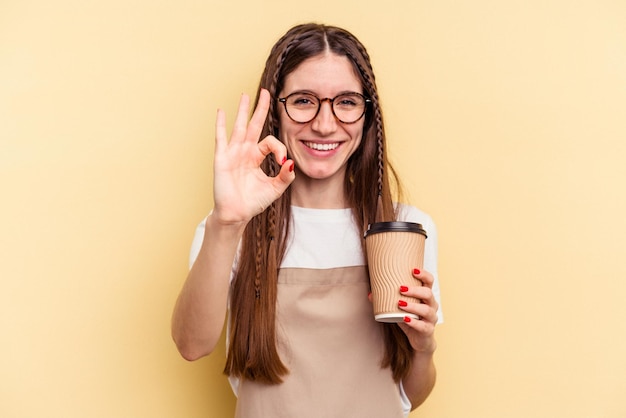 Restaurant waiter woman holding a take away coffee isolated on yellow background cheerful and confident showing ok gesture.