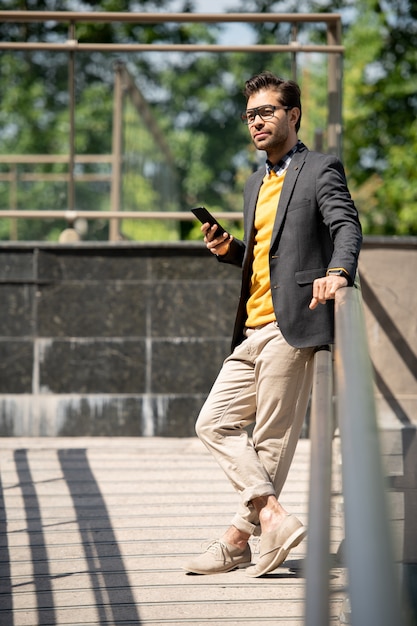 Restful young businessman in smart casual enjoying summer day in the city while scrolling in smartphone