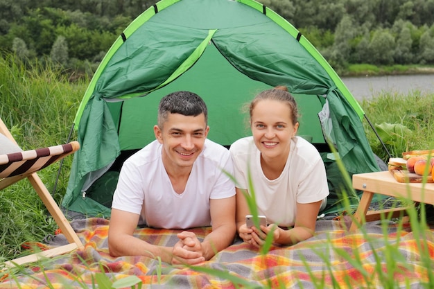 Resting outdoor Smiling woman and man wearing casual clothing laying at tent near the river looking at camera resting camping at riverside