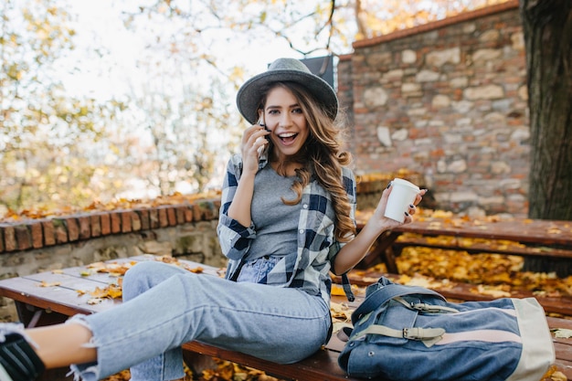 Resting white lady in short casual jeans calling someone on autumn background. Outdoor portrait of fashionable european girl in good mood enjoying coffee in november day.