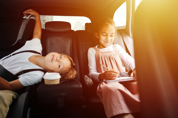 Restless siblings riding on a back seat in a car. View from inside salon. Boy is bored, he's run out of things to do. Girl is drawing.