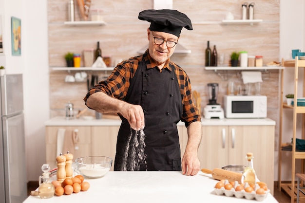 Retaried baker wearing apron and prepraing homemade pizza on kitchen table. Retired senior chef with bonete and apron, in kitchen uniform sprinkling sieving sifting ingredients by hand.