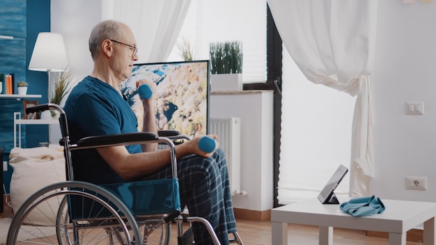 Retired adult with disability lifting dumbbells and watching video of online training program. Aged man sitting in wheelchair and following workout lesson to work arms muscles with weights