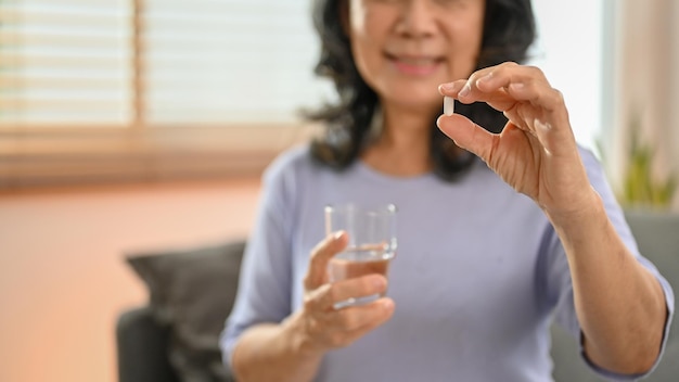 A retired Asian woman holding a glass of water and a pill taking a medicine in her living room