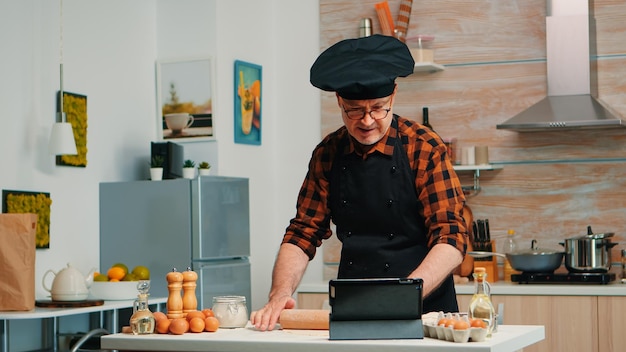 Retired man following culinary advice on tablet, learning cooking tutorial on social media, forming dough with wooden rolling pin. Grandfather with bonete and apron using laptop preparing home cakes