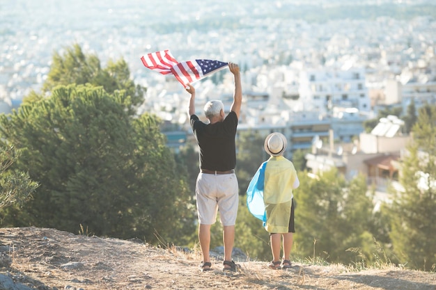 Retired man and kid boy holding the flags of the USA and Ukraine on city view