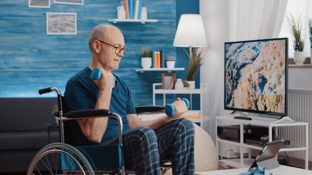 Retired man sitting in wheelchair and lifting dumbbells to exercise while watching online video of workout on tablet. Elder adult with disability using weights, following training lesson