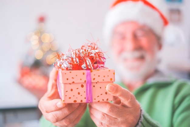 Retired senior man in santa hat and warm clothing celebrating christmas at home. Old male santa smiling while holding christmas present. Elderly man holding christmas gift box