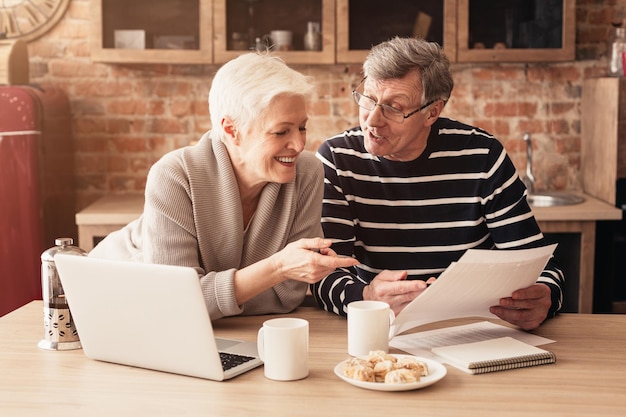 Retirement Financial Planning Concept. Happy Senior Couple Discussing Family Budget Together, Sitting In Kitchen With Laptop And Papers