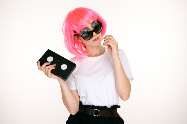 Retro girl in the glasses and pink wig holding black cassette on the white background.