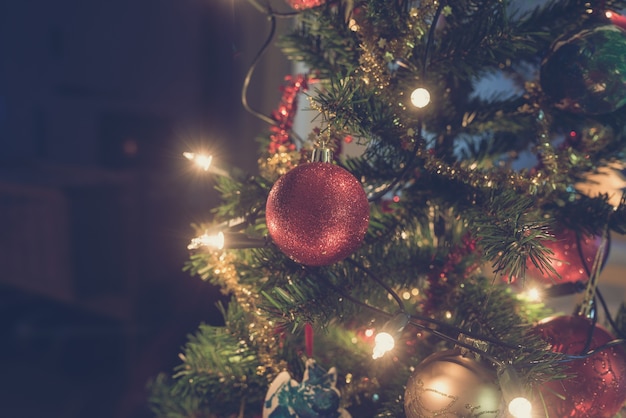 Retro image of shiny red Christmas bauble hanging on decorated holiday tree with lights.