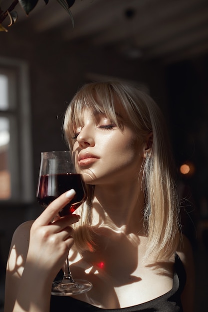 Retro portrait sexy blonde woman with wine glass in black dress, vintage interior woman posing at window. Sensual romantic look, girl relaxes at home