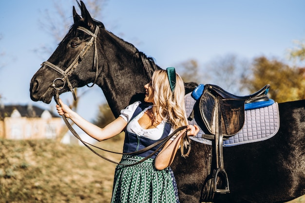 Retty blonde in traditional dress  walking with big black horse outdoors