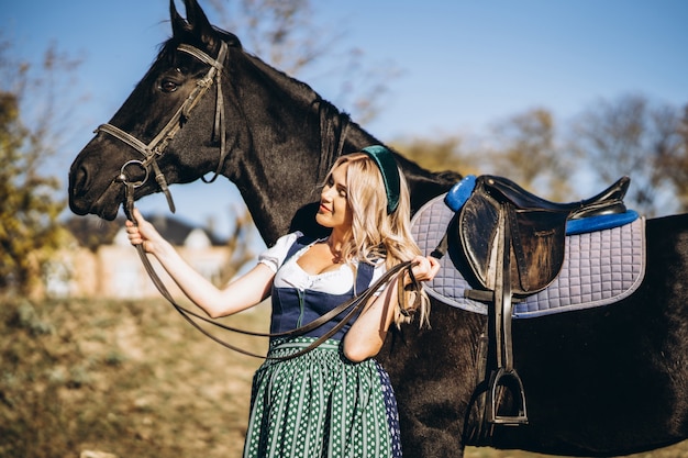 Retty blonde in traditional dress  walking with big black horse outdoors