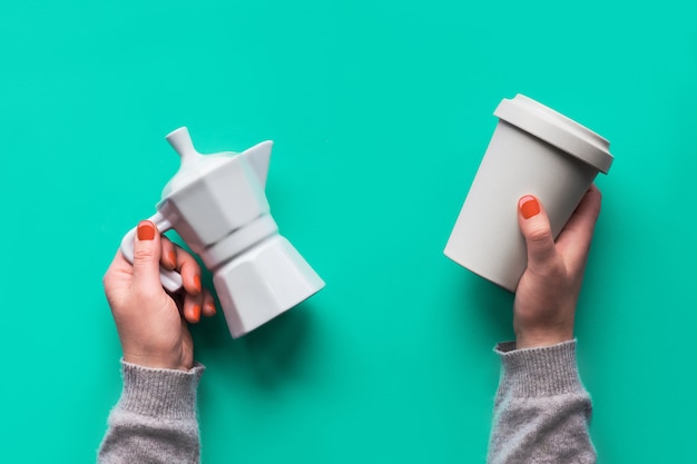 Reusable coffee mug or keep cup and white ceramic coffee maker in woman's hands on mint green wall. Creative flat lay, top view, trendy zero waste concept, reusable coffee cup with silicone lid.