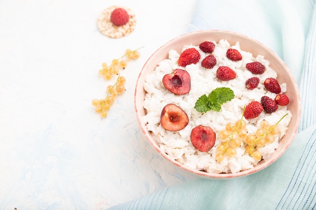 Rice flakes porridge with milk and strawberry in ceramic bowl on white concrete background and blue linen textile. Side view, close up, selective focus.