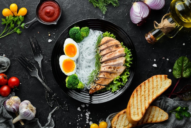 Rice noodles with chicken fillet egg and broccoli in a black plate on a black background Top view Free space for your text