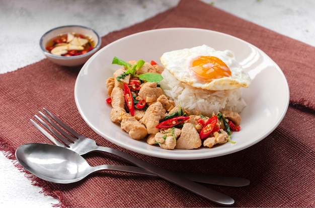 Rice topped stirfried basil with chiken and a fried egg on white plate serving on brown table cloth with fish sauce  Thai street food