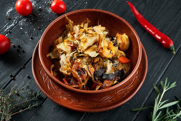 Rice wok noodles with seafood (squid chips, shrimps) soy sauce and vegetables in a ceramic bowl on black.