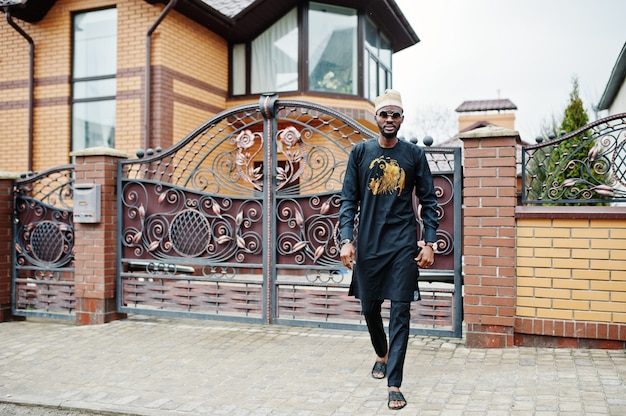 Rich african man in stylish traditional clothes and hat posed outdoor background his mansion.