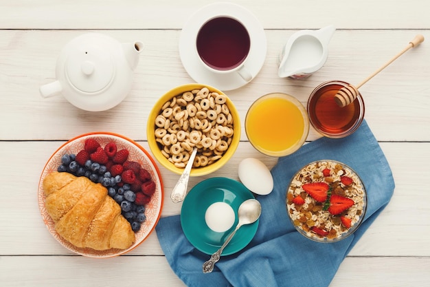 Rich breakfast menu background. French crusty croissants, muesli, glass of orange fresh, yogurt, coffee, berries, honey and boiled eggs for tasty morning meals on wooden table, top view