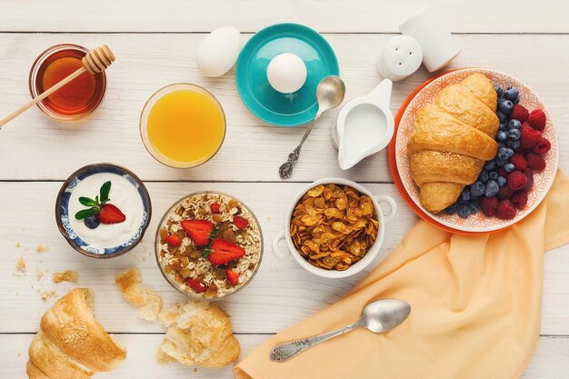 Rich breakfast menu background. French crusty croissants, muesli, glass of orange fresh, yogurt, coffee, berries, honey and boiled eggs for tasty morning meals on wooden table, top view