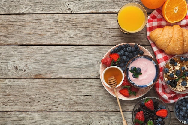 Rich tasty breakfast background. Croissants with golden crust, muesli, honey, glass of fresh orange juice and bowl of sweet organic blueberries on rustic wooden surface, top view, copy space