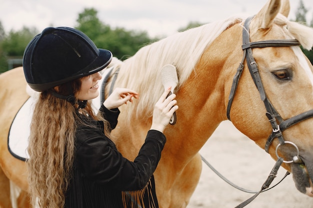 Rider woman comb her horse on a ranch. Woman has long hair and black clothes. Female equestrian touching her brown horse.