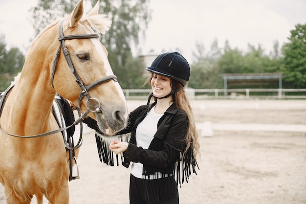 Rider woman talking to her horse on a ranch. Woman has long hair and black clothes. Female equestrian touching her brown horse.