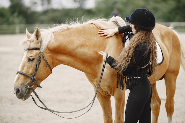 Rider woman talking to her horse on a ranch. Woman has long hair and black clothes. Female equestrian touching a saddle.