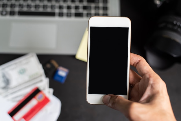 Right hand holding smartphone on blur blackground with laptop, dollars, camera, and credit card