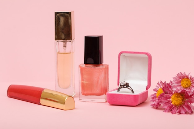 Ring in a box, nail polish, perfume, lipstick and flowers on a pink background. Women jewelry, cosmetics and accessories.