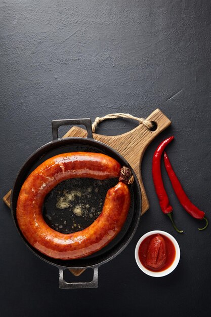 Ring of fried pork sausage on a black cast iron frying pan with hot pepper and ketchup on a black