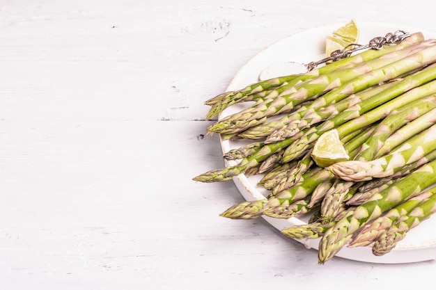 Ripe asparagus ready for cooking. Ceramic plate and lime pieces. Modern hard light, dark shadow. White wooden table, copy space