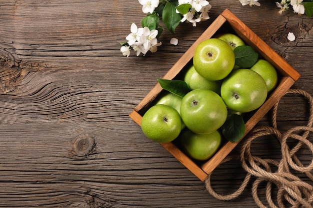 Ripe green apples with branch of white flowers in wooden box on a wooden table. Top view with space for your text.