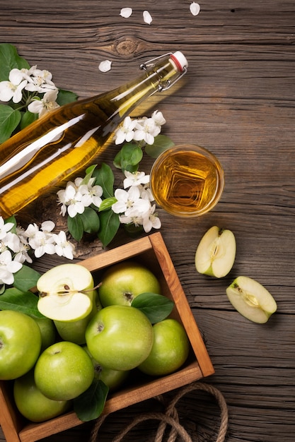 Ripe green apples in wooden box with branch of white flowers, glass and bottle of cider on a wooden table. Top view.