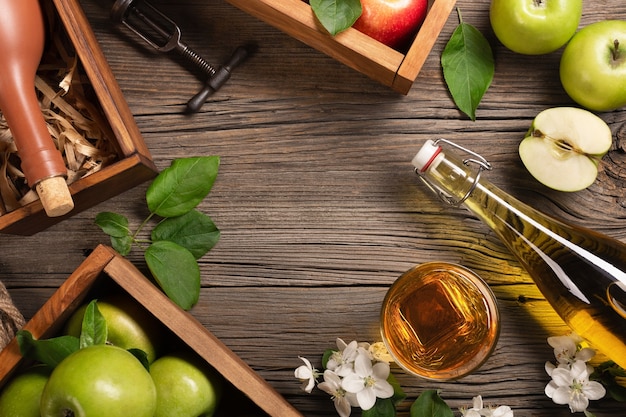 Ripe green and red apples in wooden box with branch of white flowers, glass and bottle of cider on a wooden table. Top view with space for your text.