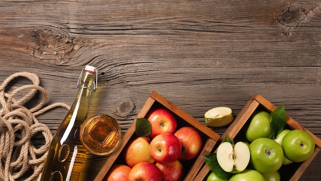 Ripe green and red apples in wooden box with branch of white flowers, glass and bottle of cider on a wooden table. Top view with space for your text.