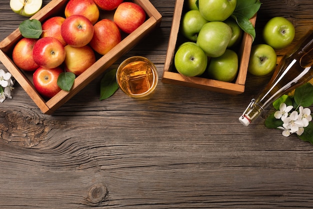 Ripe green and red apples in wooden box with branch of white flowers, glass and bottle of cider on a wooden table. Top view with space for your text.