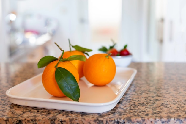 Ripe juicy oranges with leaves on a white tray on a stone table in the kitchen