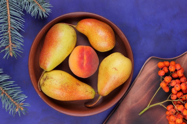 Ripe juicy pears and peach in a clay plate with fir branches on a blue background Fruits in a plate and rowanberries on a wooden board closeup