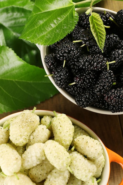 Ripe mulberries in bowls with green leaves on table close up