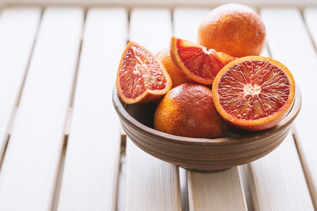 Ripe red oranges in brown bowl on white wooden background. Sliced and whole ripe juicy Sicilian Blood oranges.