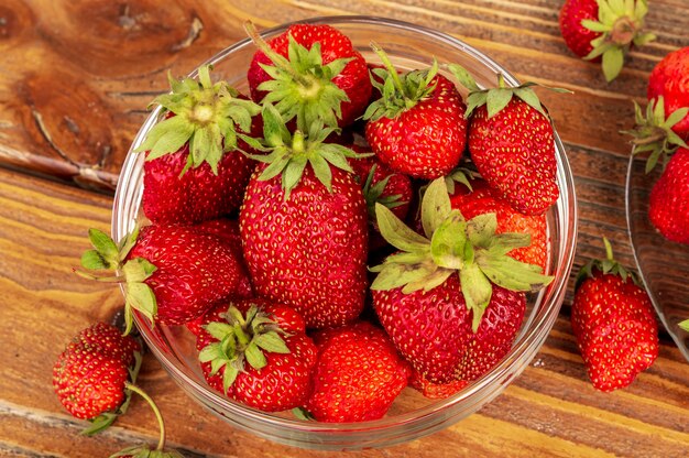 Ripe red organic strawberries in a glass bowl against brown wooden background.