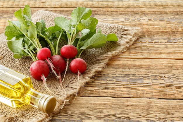 Ripe red radishes with green stalks and a bottle of oil on sackcloth and old wooden boards. Top view. Spring harvesting of vegetables in a garden.