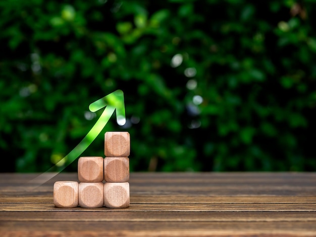 Rise up glowing arrow on empty wooden cube blocks as a business chart steps on table and green leaves background with copy space Business growth investment sustainable development goals concepts