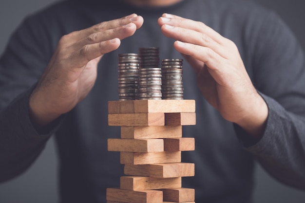 Risks in business or financial concept Idea to prevent risk in business Business man using hand to protection coin stacked on tower wooden block game and prevent falling down