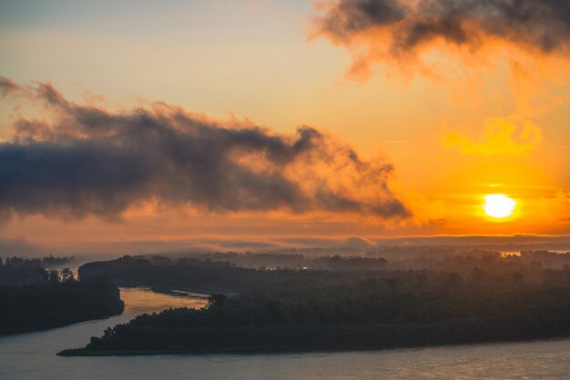 River flows along shore with forest. Channel flows around island in fog. Orange glow around sun at dawn in cloudy sky reflected on water. Morning atmospheric landscape of majestic nature in backlight.