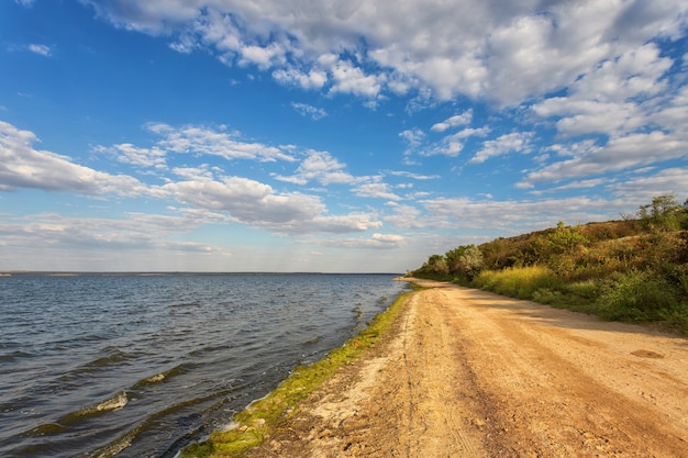 The road along the shore of the lake, river, against a blue sky with belvmi clouds