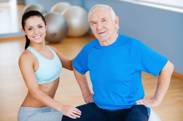 On the road to full recovery. Confident female physical therapist looking at camera and smiling while sitting close to cheerful senior man